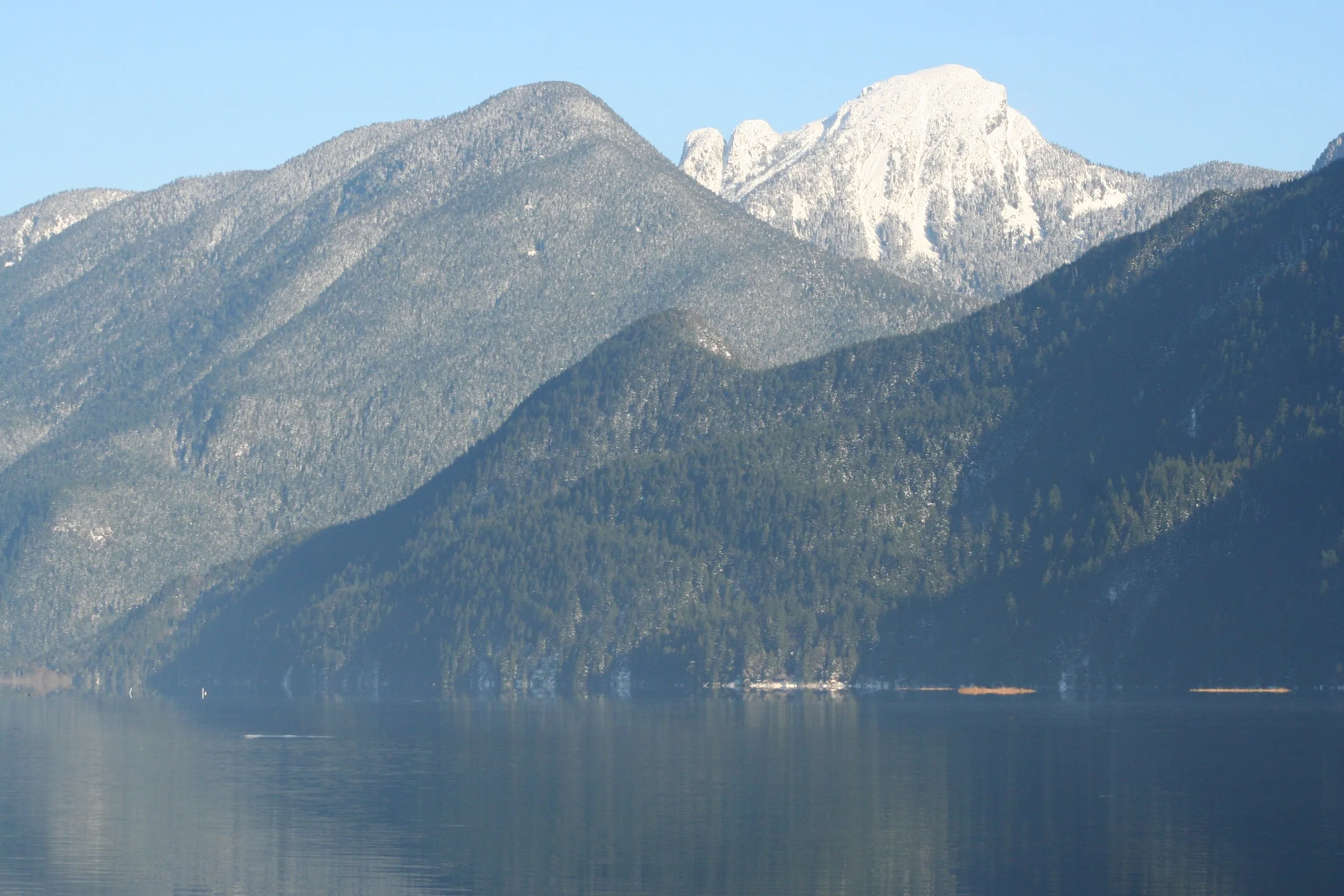 Snow dusted mountains rising above the calm water of Pitt Lake under a clear blue sky.