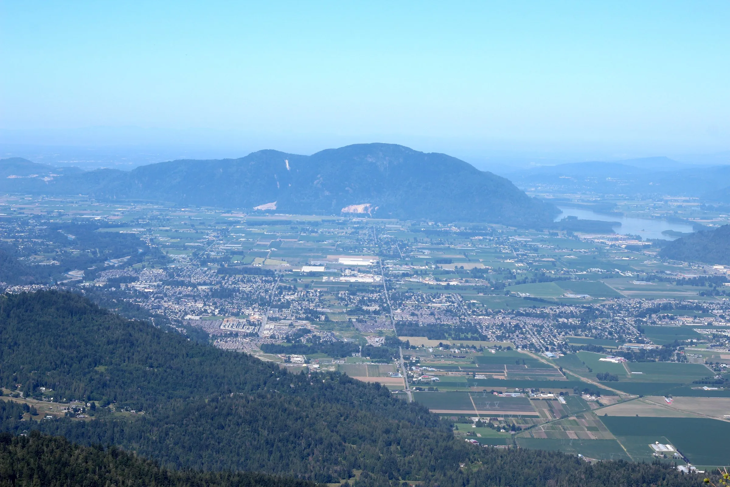 A wide view over Chilliwack and its farmland from a forested mountainside with a mountain and river in the distance.