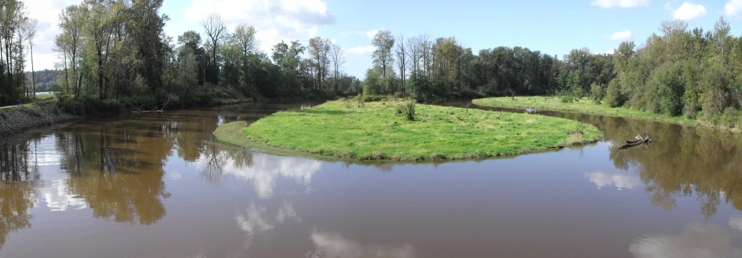 Kanaka Creek curving around a grassy island under a blue sky with trees reflected in the water in Maple Ridge.