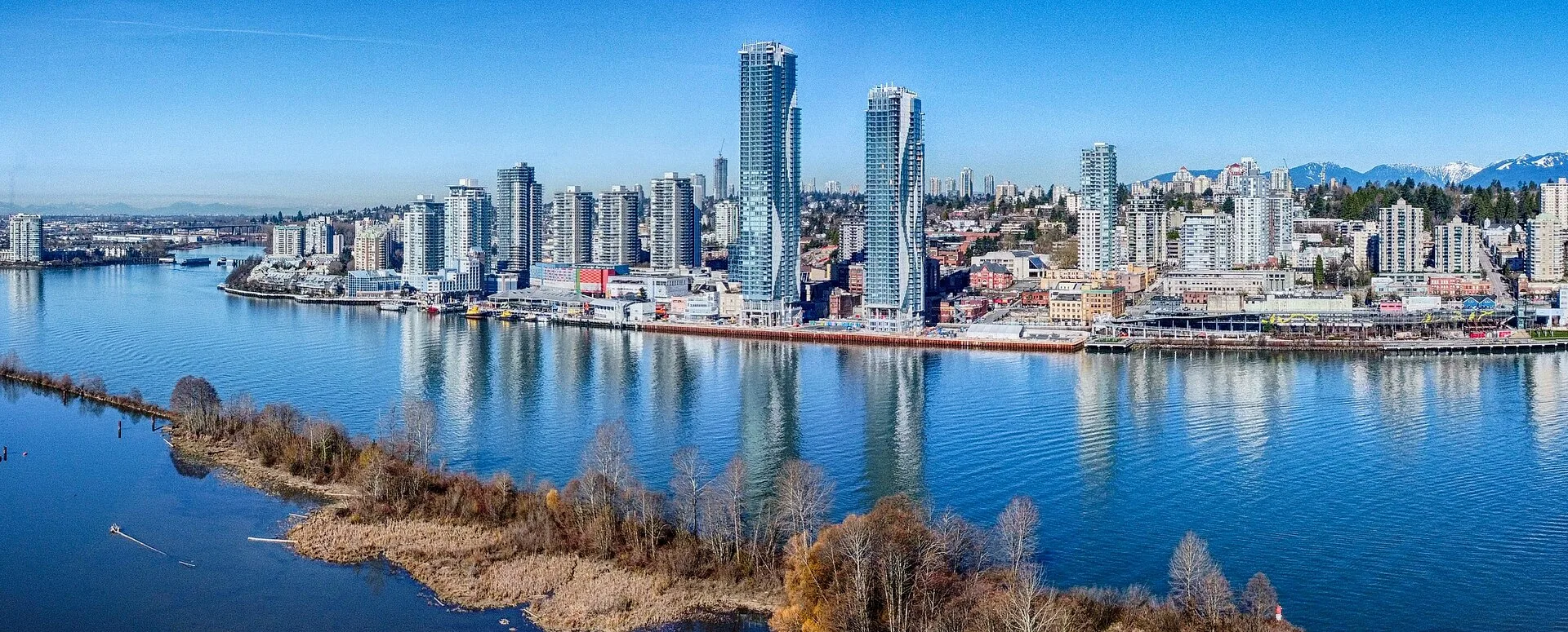 The New Westminster waterfront towers along the Fraser River, seen from across the water with mountains in the distance.