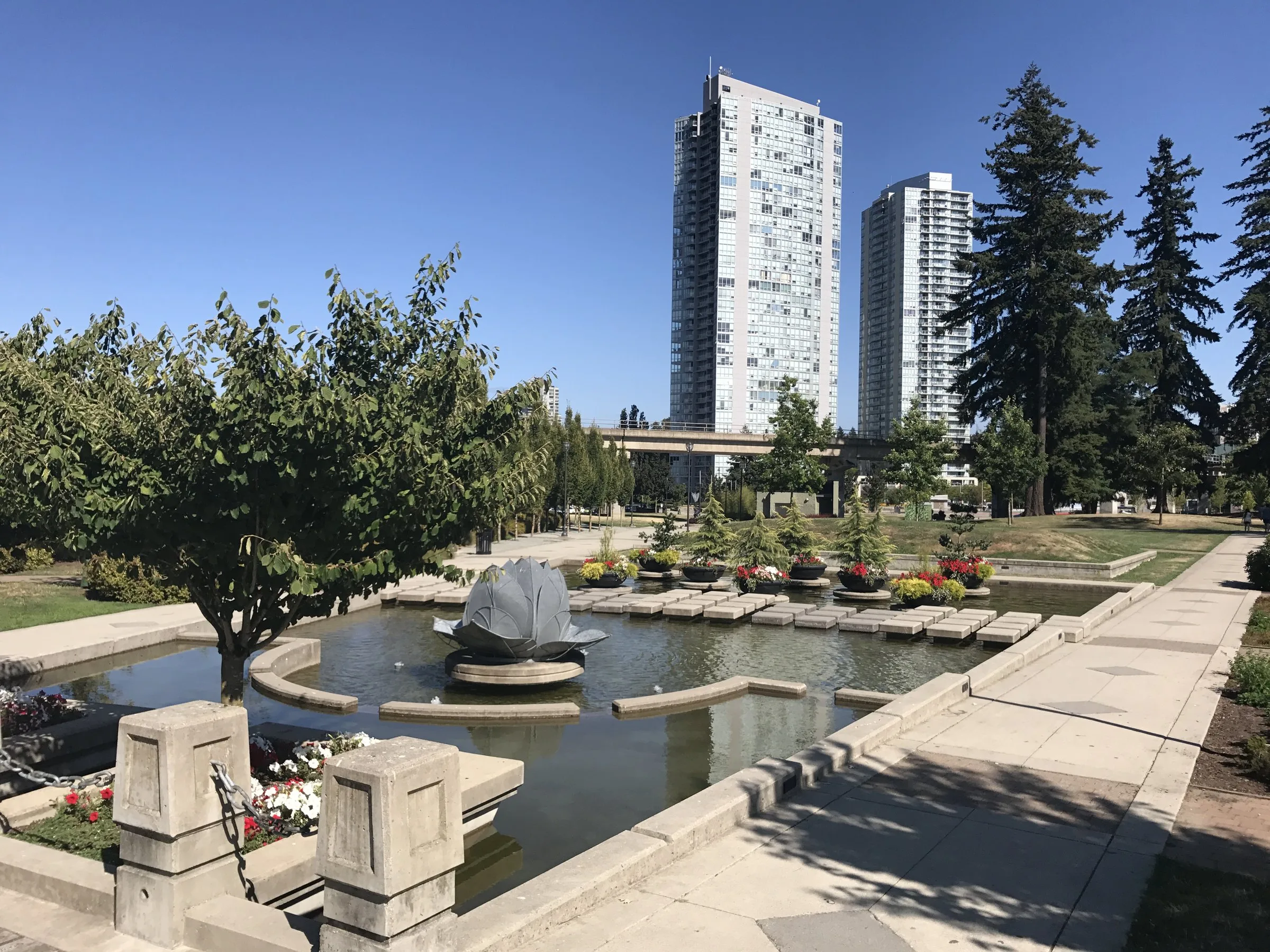 A fountain pool and flower beds in Holland Park with Surrey City Centre towers behind under a clear sky.